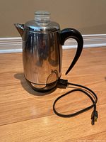 Side view of the stainless steel coffee percolator with black handle and electric cord