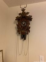 Full front view of the cuckoo clock hanging on a wall showing overall size and shape with hanging weights and chains.