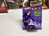 Unopened 2014 Hot Wheels Batman Catwoman themed die-cast car in original packaging displayed on white surface with background items.