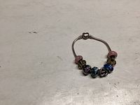 Full view of silver charm bracelet with 8 assorted charms including pink glass beads, blue enamel, silver ornate charms on white background.