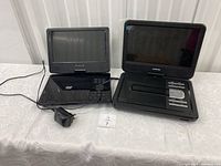 Two black portable DVD players on a white table, one with charger connected and one without charger. Both with foldable screens and control buttons visible.