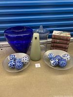 Full lot of decor items including blue glass vase, glass candy dish, green candle, porcelain balls in bowls, and faux book storage boxes.