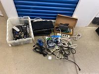 Full lot view showing plastic bin with remote controls, stacked electronics including receivers and Blu-ray player, assorted cables and cords on floor, and power strip.