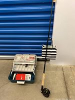 Photo of fly fishing rod standing upright beside a fishing tackle box on a concrete floor. Shows overall length and handle detail.