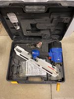 Open black hard plastic carrying case containing a silver and blue framing nailer, some instructions, and small parts.