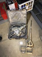 Full lot with Makita electric drill in plastic case and two metal mixing paddles on concrete floor.