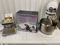 White stand mixer with whisk and bowl on right, boxed Vitantonio Pizzelle Baker waffle maker in center and chrome Atlas pasta maker with yellow handles on left