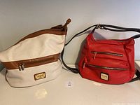 Cream white leather handbag with brown trim and single shoulder strap next to red leather handbag/backpack with multiple zipper pockets, on display against neutral background