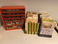 Orange storage organizer with multiple drawers containing various sewing notions, buttons, and thread. Next to two boxes holding a large number of assorted sewing patterns in their original envelopes.