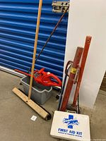 Photo showing red electric shrub trimmer, push broom with wooden handle, gray bucket, orange fence post hammer, and sledgehammer leaning against white wall.