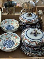 Porcelain bowls and plates with floral patterns, lidded serving pots with ornate handles, and silver-tone metal cups with handles.