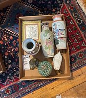 Box with three Asian vases, two Buddha figurines, snail figurine, and framed wall art on a patterned carpet