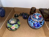 Several enamelled jars and one decorated metal cup arranged in a cardboard box showing variety of floral designs and colorful patterns.