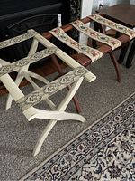 Photo showing two wooden luggage racks side by side, one white with decorative green and red patterned straps, and one natural wood with floral embroidered straps.