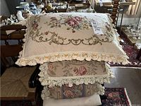 Stack of four hand cross-stitched pillows with floral designs, decorative tassel trim, showing front needlepoint sides and velveteen backs underneath.