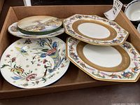 Five decorative plates in cardboard box, viewed from above. Includes various shapes and designs: bird motifs, floral, and rustic scenes.