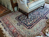 Full view of the large floral pattern area rug under a sofa, showing intricate design and color scheme, including navy blue center, red and light blue borders, and fringed edges.