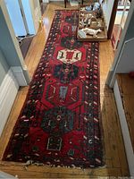Long view of antique runner rug on hardwood floor with visible wear and frayed edges. Shows geometric hexagonal and linear patterns in red, blue, black, and white.
