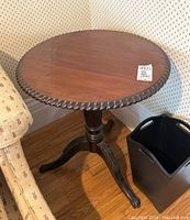 Full view of round wooden side table next to a floral upholstered chair and black basket on hardwood floor