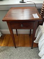 Front view of smaller wood side table with drawer and round metal knob on four tapered legs on wood floor with carpet