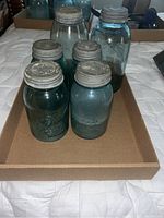 Six vintage Ball mason jars with zinc lids in a cardboard tray on a bed. The jars show blue-green tint and have worn, dirty zinc lids.
