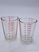 Two vintage glass measuring cups, showing red measurement marks in ml, oz, and tablespoons.