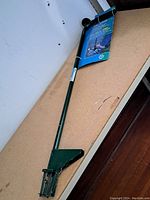 Full view of the green metal heavy duty garden weeder with curved handle and digging tines at the end, showing the entire tool resting on a wood-colored surface.