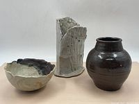 Three handmade clay pottery pieces displayed together: a dark brown round vase, a tall textured vase, and a scalloped-edge bowl.