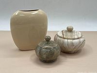 Photo showing two stone lidded bowls and a beige ceramic vase for size and style comparison.