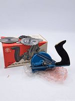 Blue metal universal saw and slotting attachment with black handle placed in front of its original box. Three circular saw blades shown on top of the box.