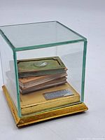 Photo of vintage square glass display case containing a stack of four miniature wooden books on a gold-colored base.