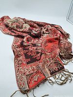 Overall view of the rectangular textile showcasing the woven floral and ornamental pattern in red, black, beige, and tan colors with tassels on the edges.