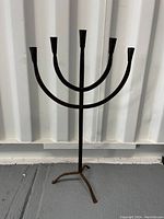 Front view of vintage wrought iron candelabra showing five candle holders and tripod base on a gray floor against a metal wall.