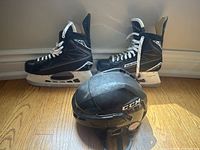 Two black Bauer Supreme hockey skates size 9EE with white laces, with a black CCM hockey helmet placed in front on a wood floor.