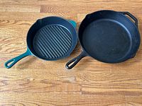 Two cast iron skillets side by side on wood floor. Left: green handled grill pan, right: black smooth skillet.