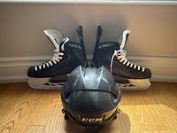 Front view of black CCM Ribcore hockey skates with white laces and black interior, positioned behind a black CCM hockey helmet. The hardwood floor background highlights the condition and details of the items.