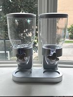 Front view of the dual cereal dispenser showing two clear containers with silver lids and turn knobs on a silver base in front of a window.