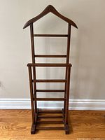 Front view of wooden clothing valet stand showing hanger-shaped top, multiple bars, and slatted base on wooden floor against white wall.