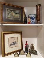 First photo showing two framed artworks on shelves along with various vases and statues, including a tall brass vase, a blue and white porcelain vase, and small decorative statues.