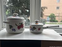 Two enamel pots from the Freitas Collection displayed side by side on a white surface in front of a window. Larger pot has two side handles, smaller pot has a long handle. Both are decorated with fruit patterns.