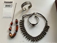 Photo of three ethnic jewelry pieces on a white background: silver cuff bracelet with wavy design, silver and fabric necklace with silver teardrop charms, and silver cuff with agate stone.