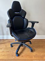 Front-left angled view of black gaming chair with blue trim on a wooden floor against a white wall