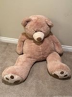 Front view of large light brown stuffed teddy bear sitting against wall on carpeted floor showing full body, face, and paws.