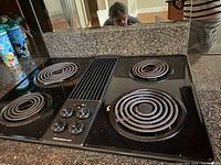 Black Jenn-Air electric inset stove top with four coil burners, control knobs on left, and vent area in the center, placed on countertop with reflective backsplash.