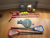 Top view of sports equipment showing tennis balls, baseball, volleyball, colorful Nike soccer ball, three pink lacrosse sticks, two pickleball paddles (Jameson and red), and a tube of tennis balls.