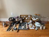 Kitchenware lot laid on wooden floor against wall including pots, pans, mugs, ceramic ware, utensils and storage jars.