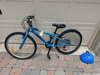 Blue Miele kids bike side view with black adjustable seat, tires, and attached reflectors, set against a garage door and brick pavement, accompanied by a blue helmet.