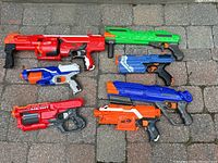 Seven various Nerf toy blasters arranged on pavement, showing colors and relative sizes.