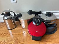 Full view of all items in the lot showing kettle, pressure cooker, red Oster waffle maker, silver Oster waffle maker, and baking dish.