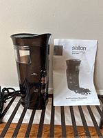 Salton smart coffee grinder standing upright with black body, clear bean compartment, and original instruction booklet beside it on a wooden slatted surface against a plain wall.